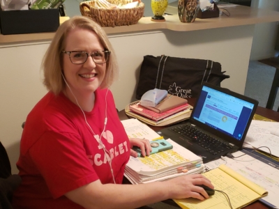 Eryn Ruder sits in a makeshift office in her kitchen, surrounded by papers and a laptop. She is wearing a red shirt and smiling.